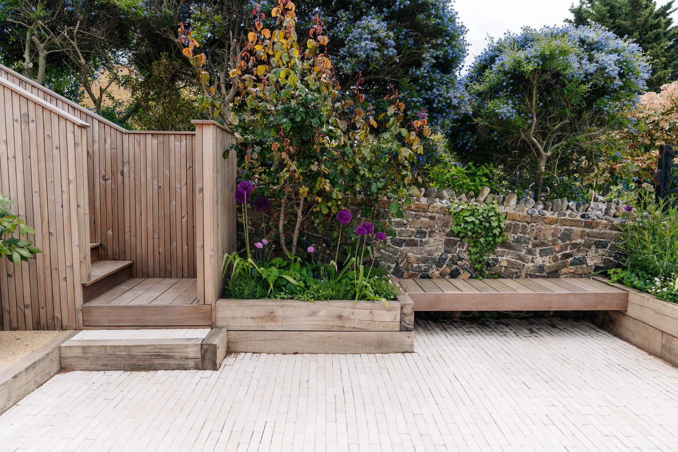 Clay-paver terrace with a built-in timber bench, sleeper steps and raised beds against a flint and brick wall