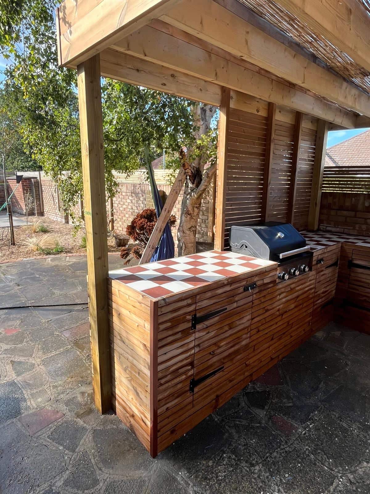 Timber outdoor kitchen under a pergola with a red and white tiled worktop, slatted storage doors and a built-in barbecue