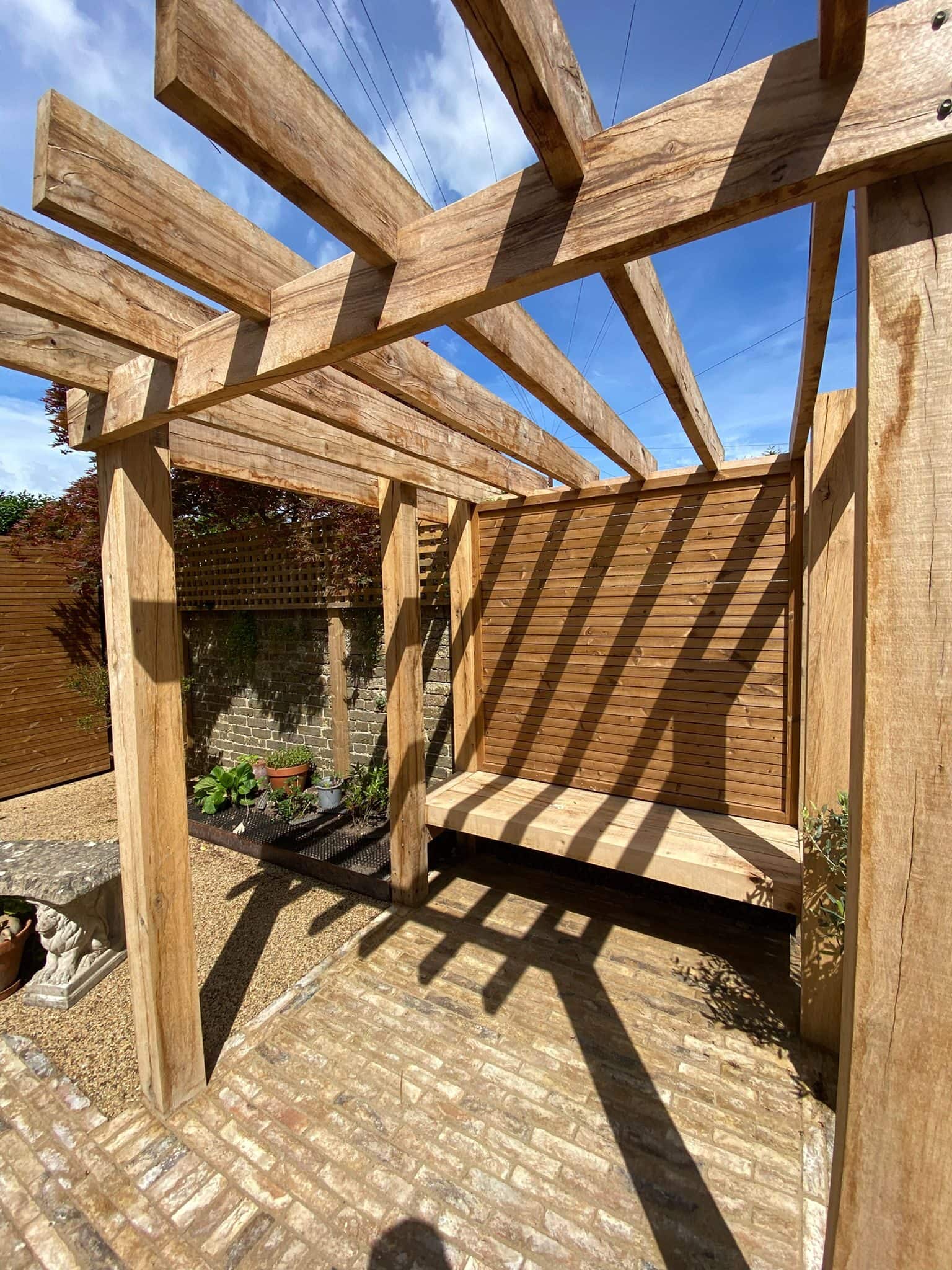 Oak pergola casting dappled shadows over a clay-paver patio and built-in timber bench